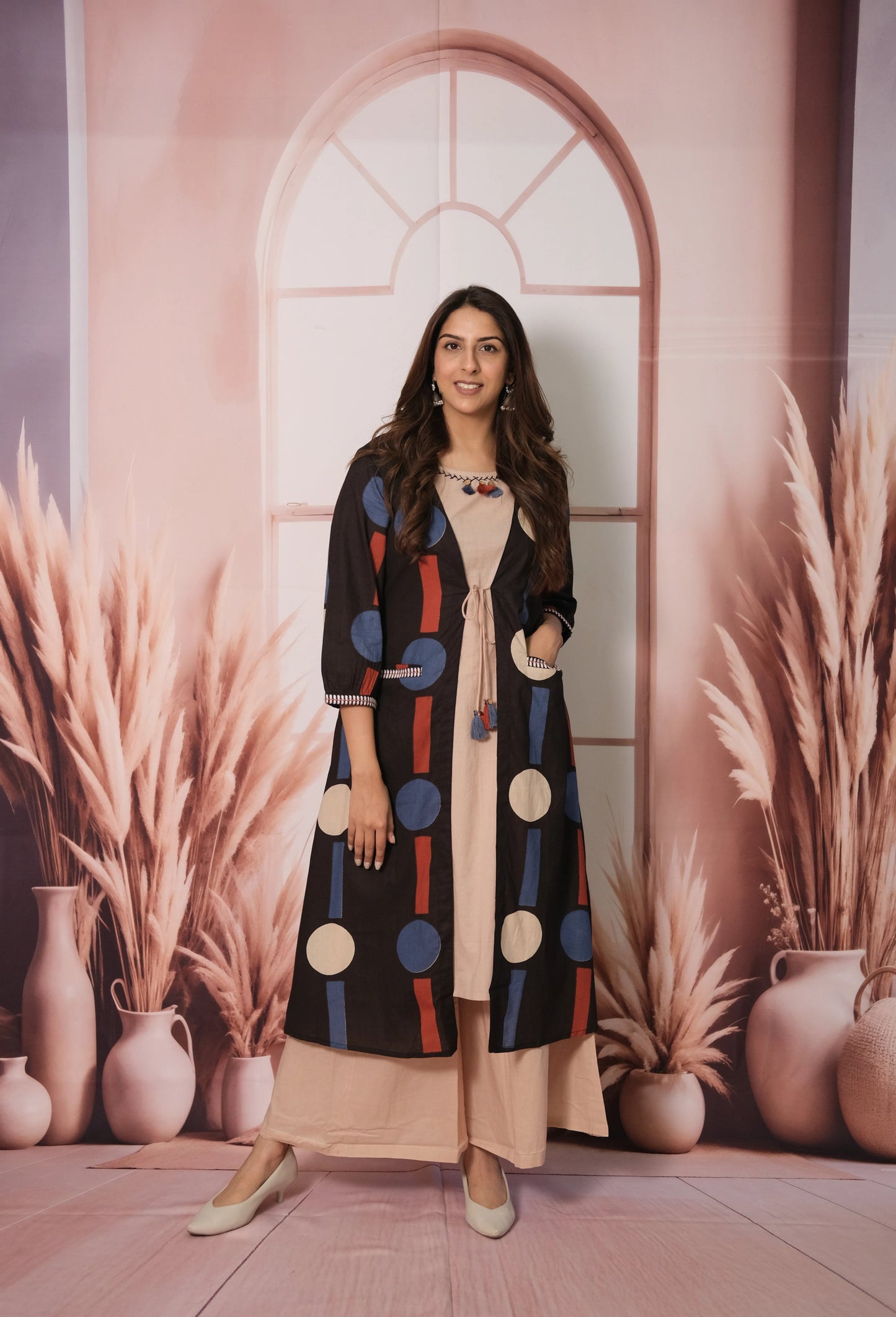 Woman wearing a colorful polka dot coat standing in front of a decorative wall with vases and plants.