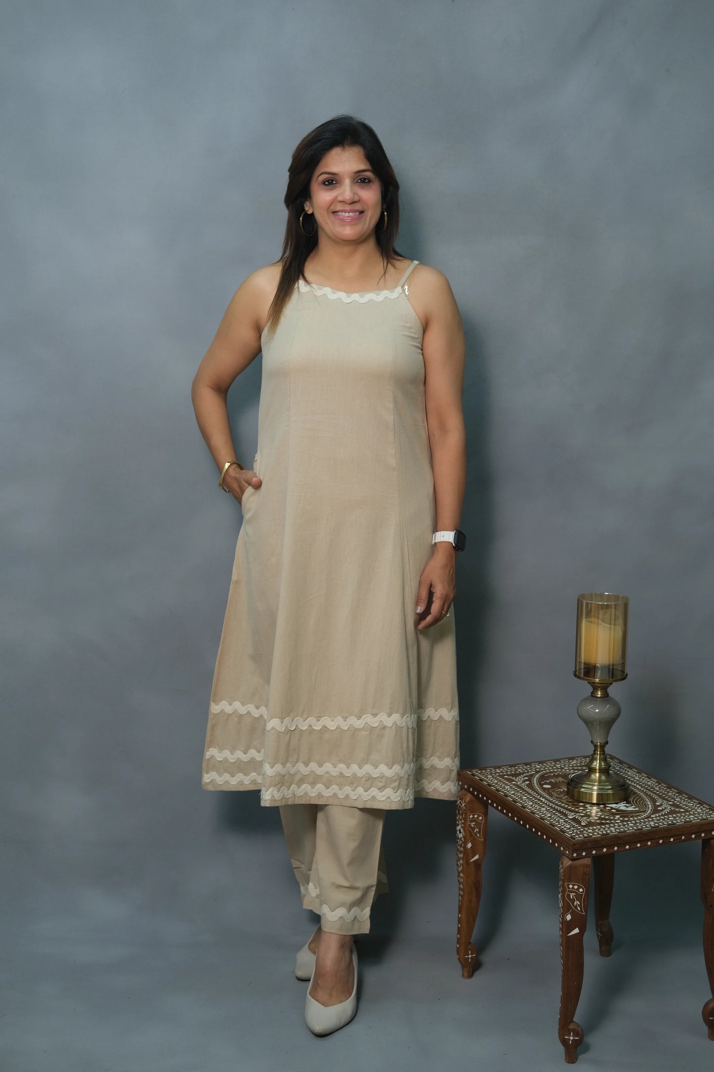 A woman stands confidently in a beige kurta with delicate white trim, paired with matching pants, and elegant heels beside a decorative candle holder.