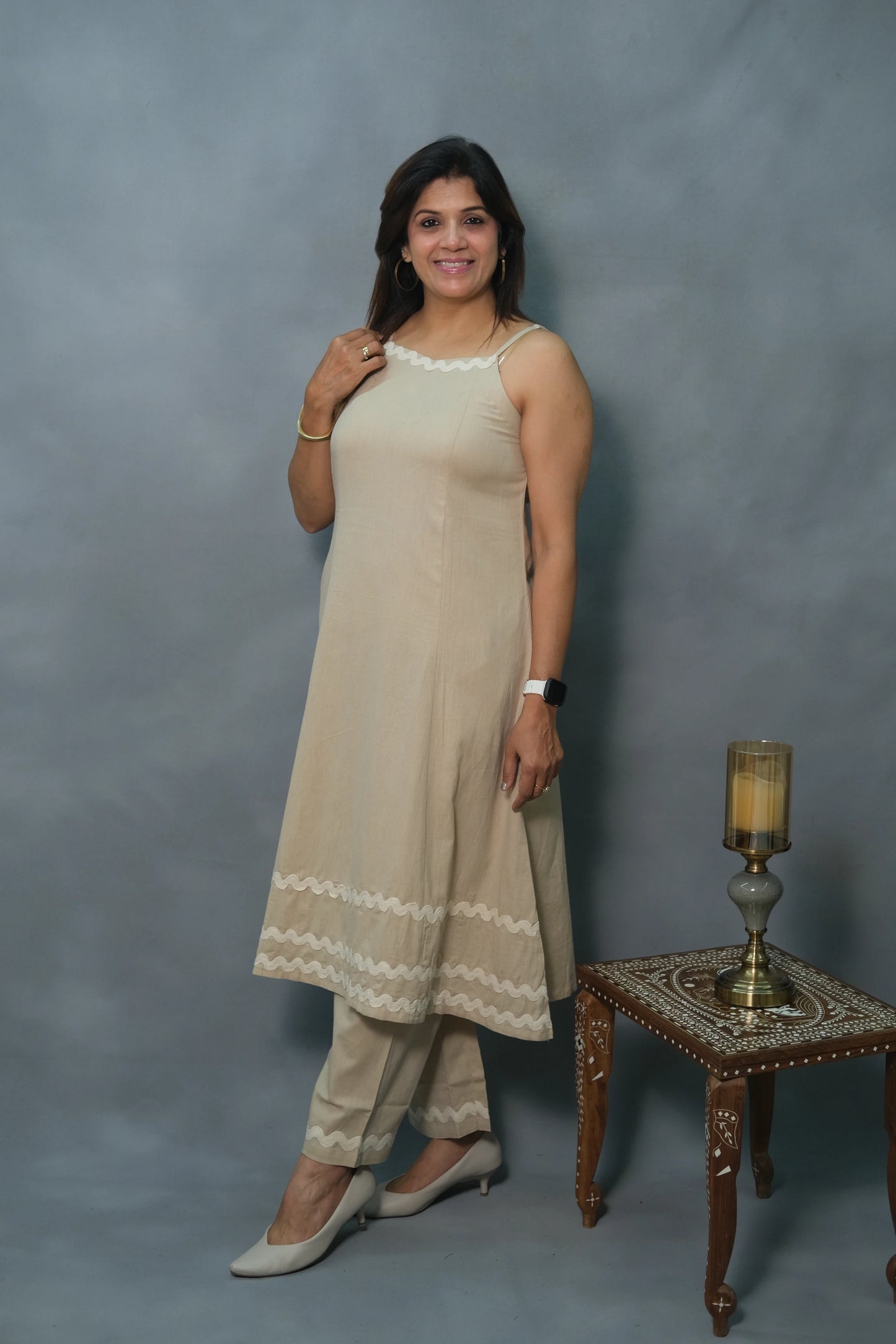 A woman in a beige ethnic outfit stands next to an ornate table with a candle holder, showcasing elegance against a gray backdrop.