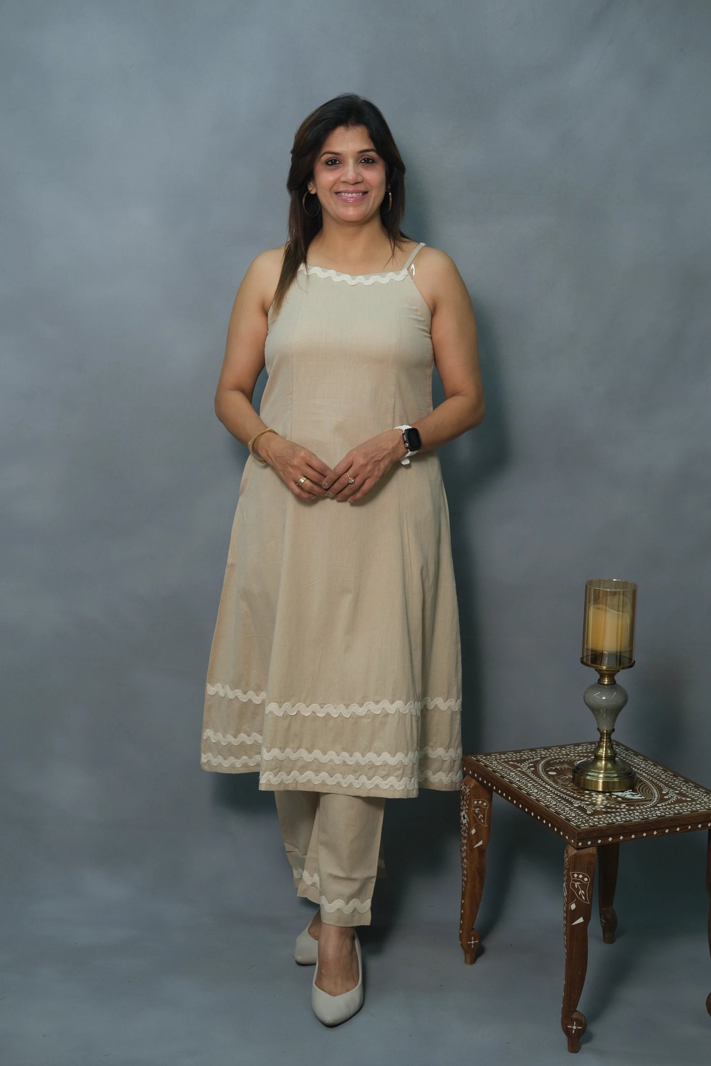 A woman stands confidently in a light beige outfit with wavy trim, paired with cream heels and stylish jewelry, beside an ornate table.