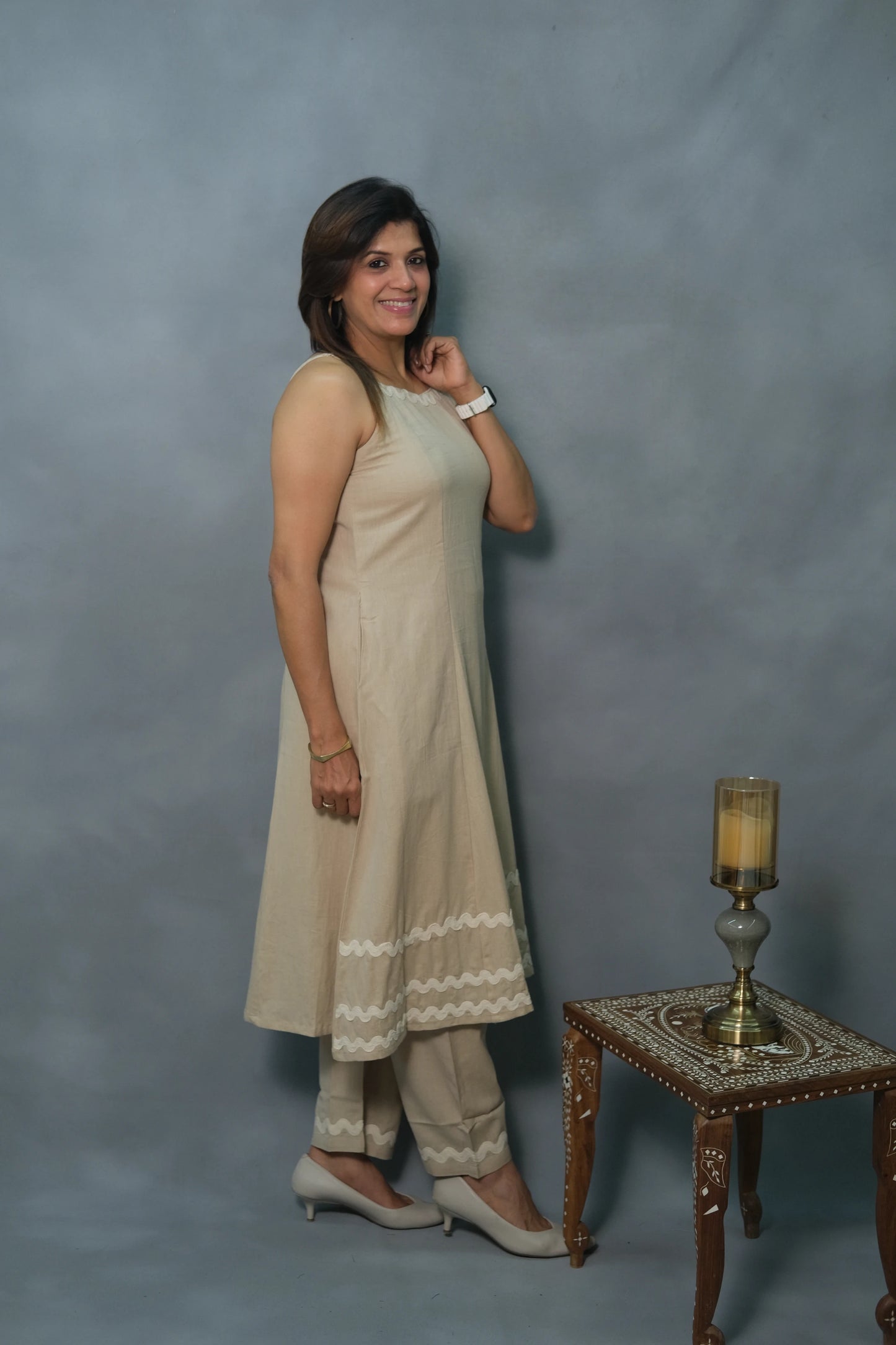 A woman in a beige ethnic outfit poses beside an ornate wooden table with a decorative candle holder, set against a gray backdrop.