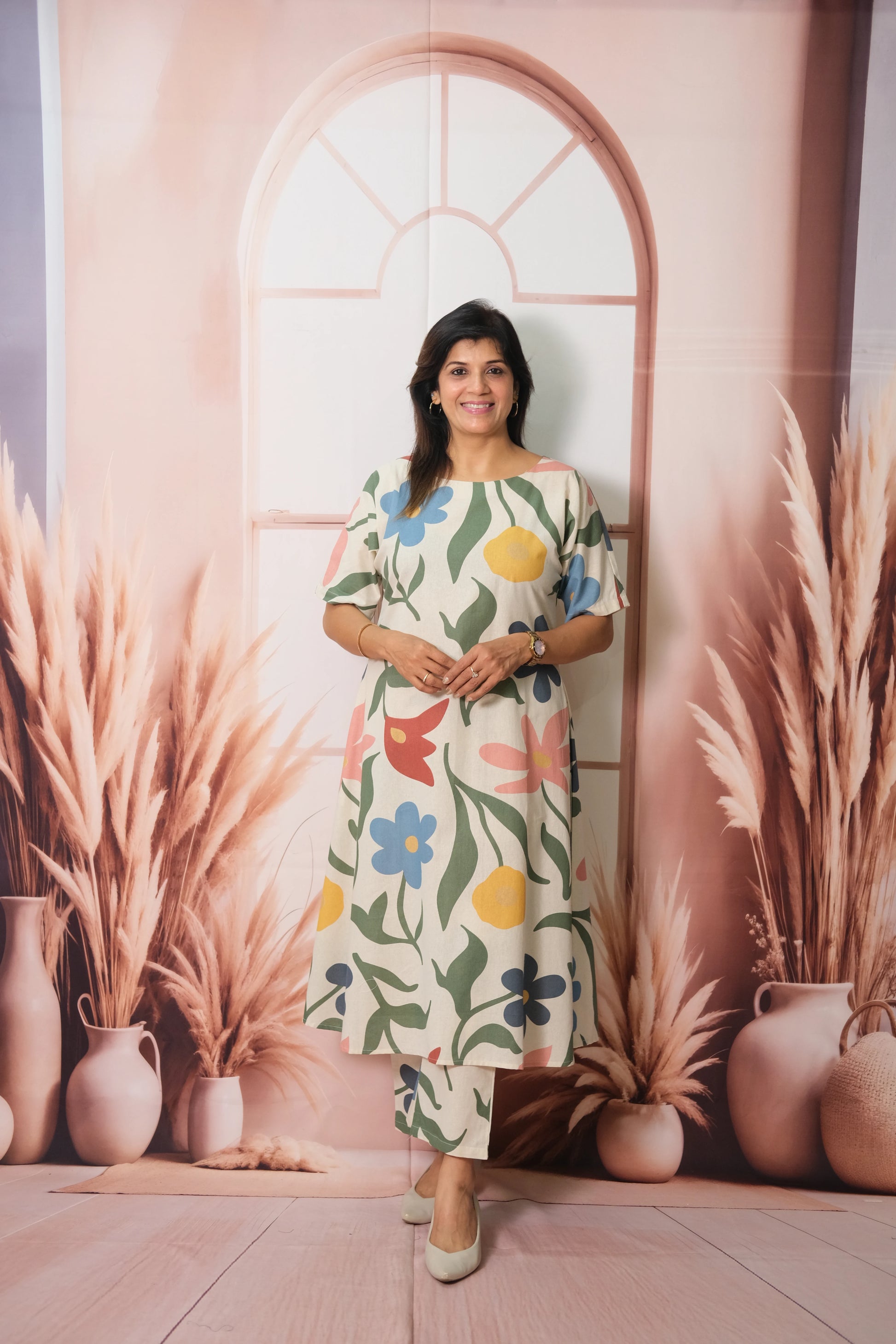 Woman in a floral dress standing in front of a decorative wall with vases and plants.