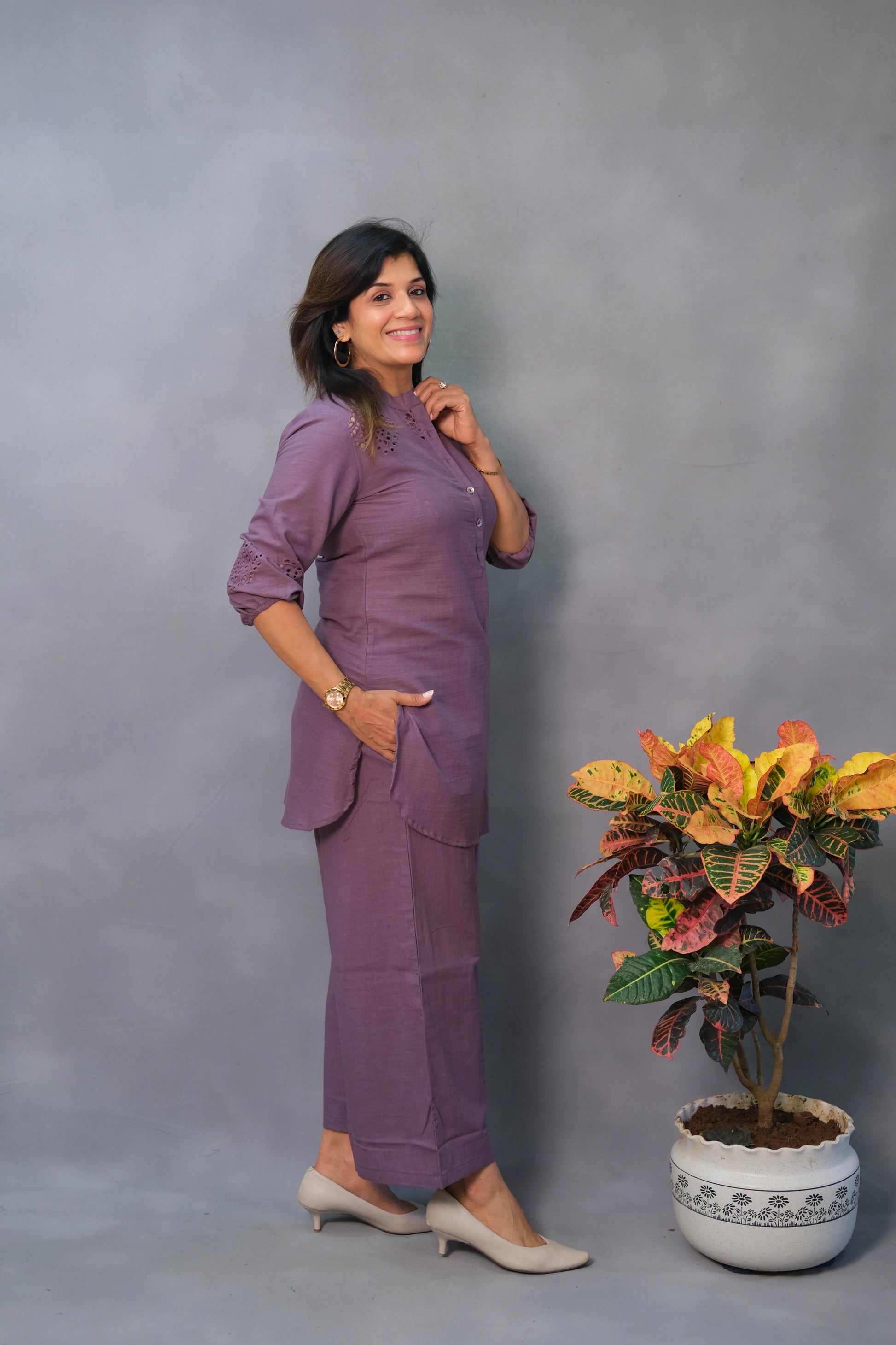 Woman in a purple outfit standing next to a potted plant against a gray background