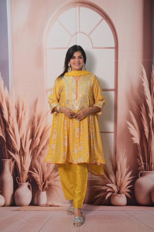 Woman in a yellow traditional outfit standing in front of a decorative wall with plants.