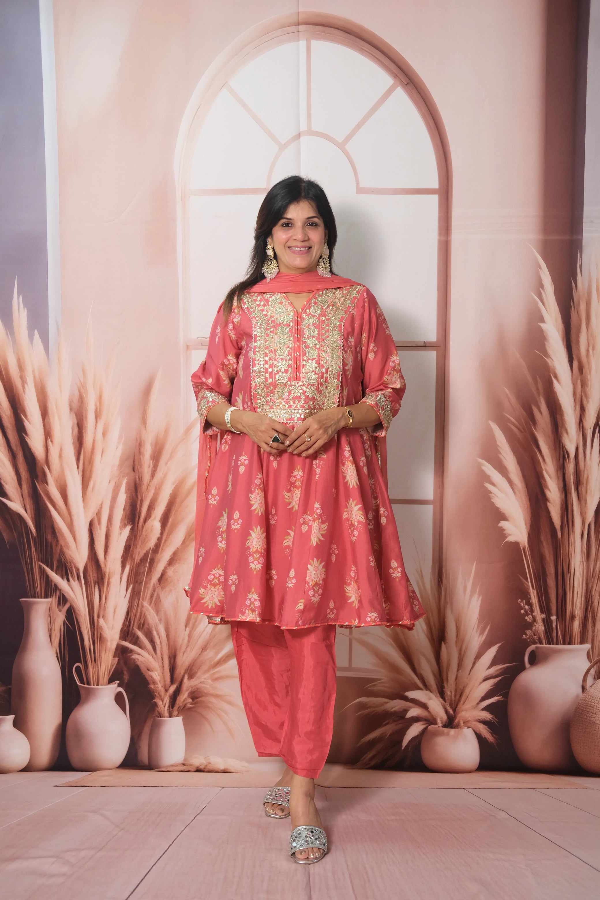 Woman in a pink traditional outfit standing in front of decorative plants and vases.