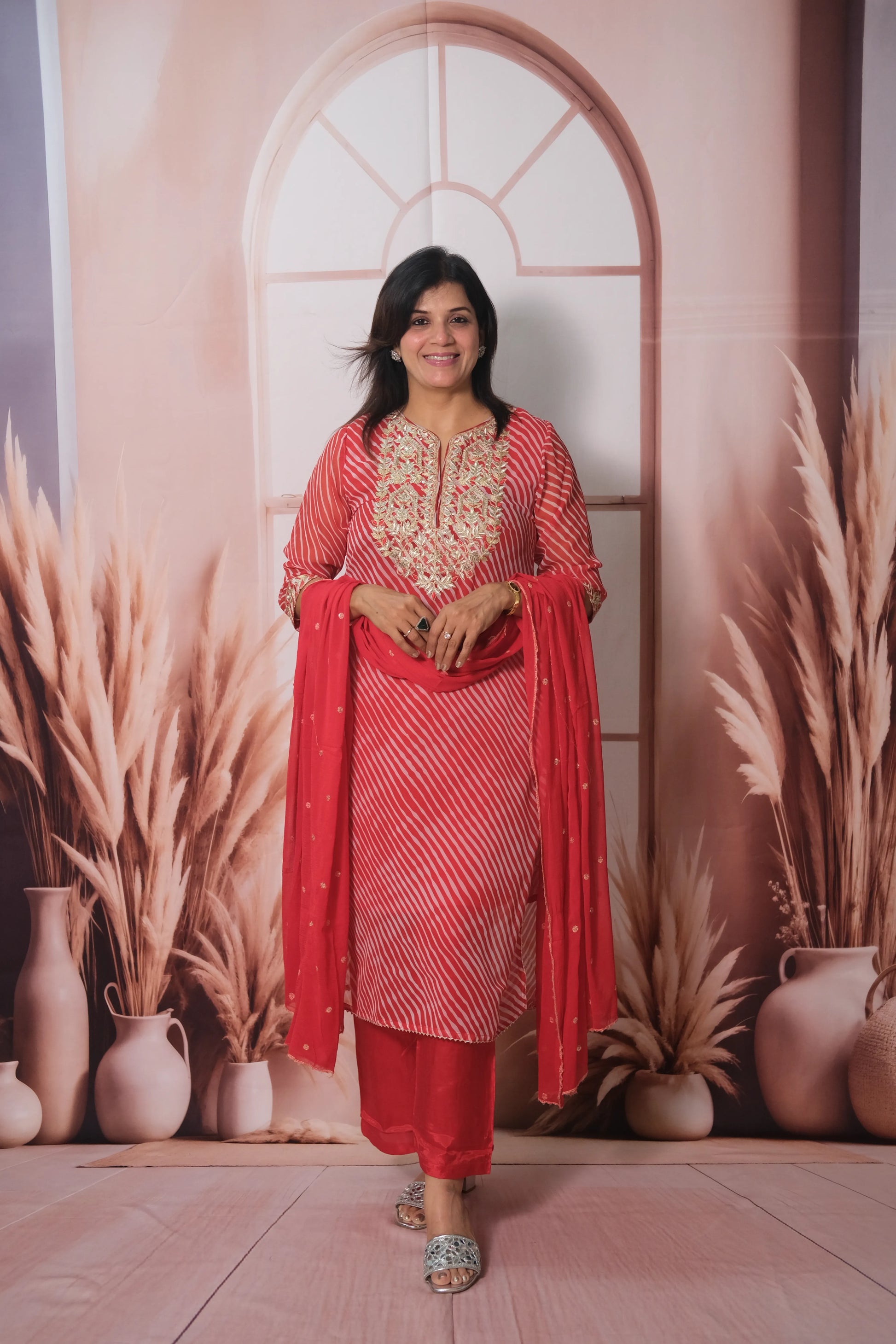 Woman in a red traditional outfit standing in front of decorative archway and plants.