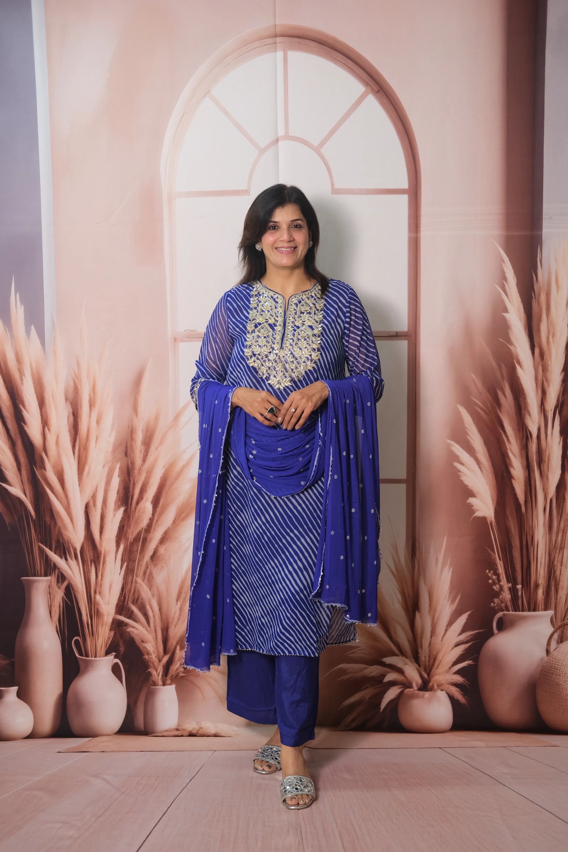 Woman in a blue traditional outfit standing against a decorative wall with arches and plants.