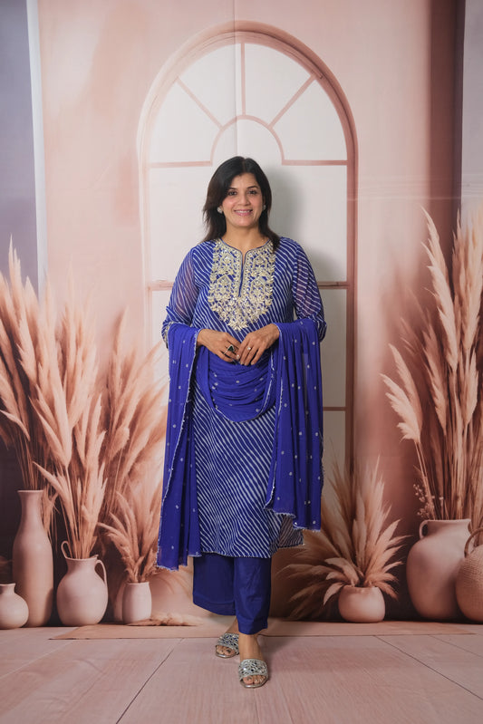 Woman in a blue traditional outfit standing against a decorative wall with arches and plants.