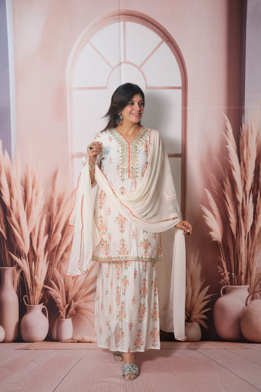Woman in a white floral dress standing in front of a decorative wall with plants and vases.
