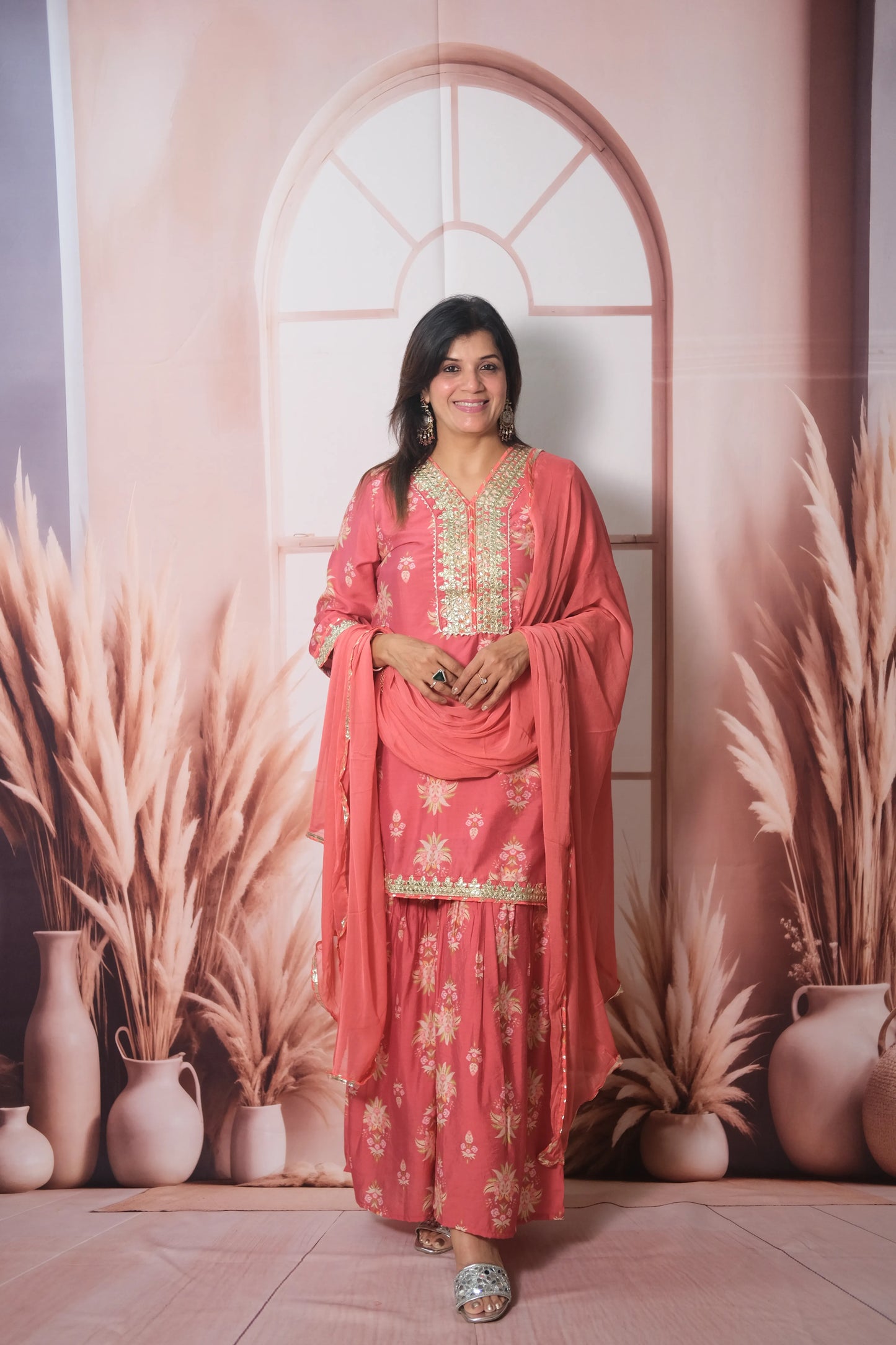 Woman in a pink traditional outfit standing against a decorative wall with plants and vases.
