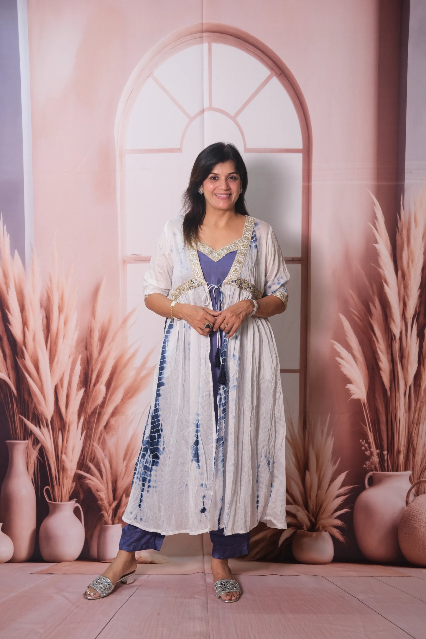 Woman in a long, patterned dress standing against a decorative wall with arches and plants.