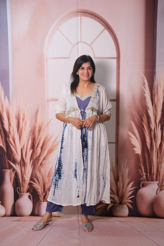 Woman in a long, patterned dress standing against a decorative wall with arches and plants.