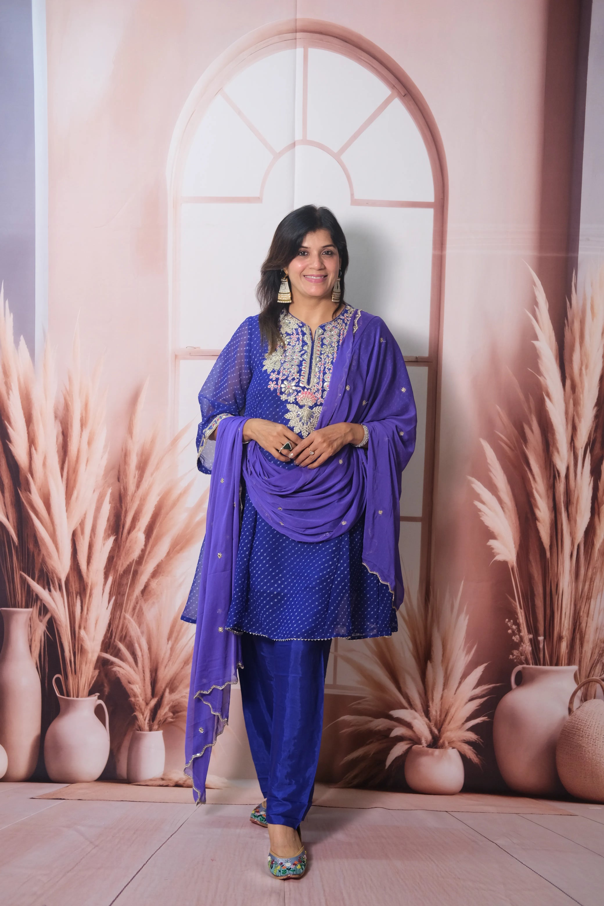 Woman in a blue traditional outfit standing against a decorative wall with arches and plants.
