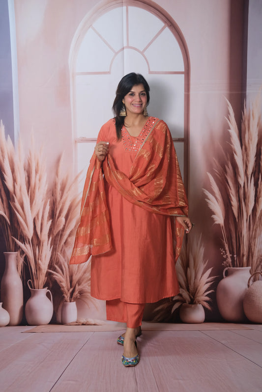Woman in an orange traditional outfit standing against a decorative wall with plants.