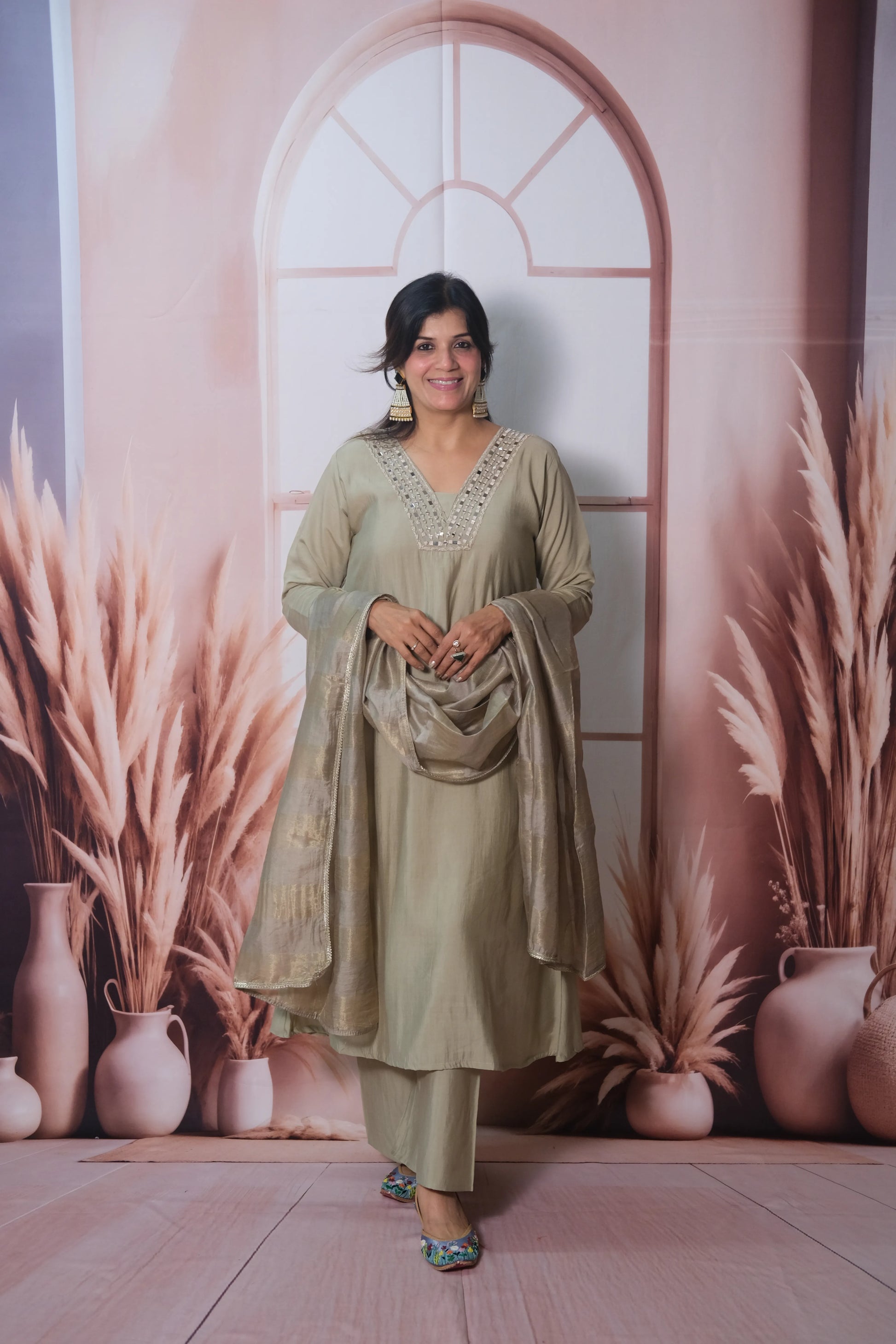 Woman in a gray traditional outfit standing in front of a decorative wall with plants and vases.