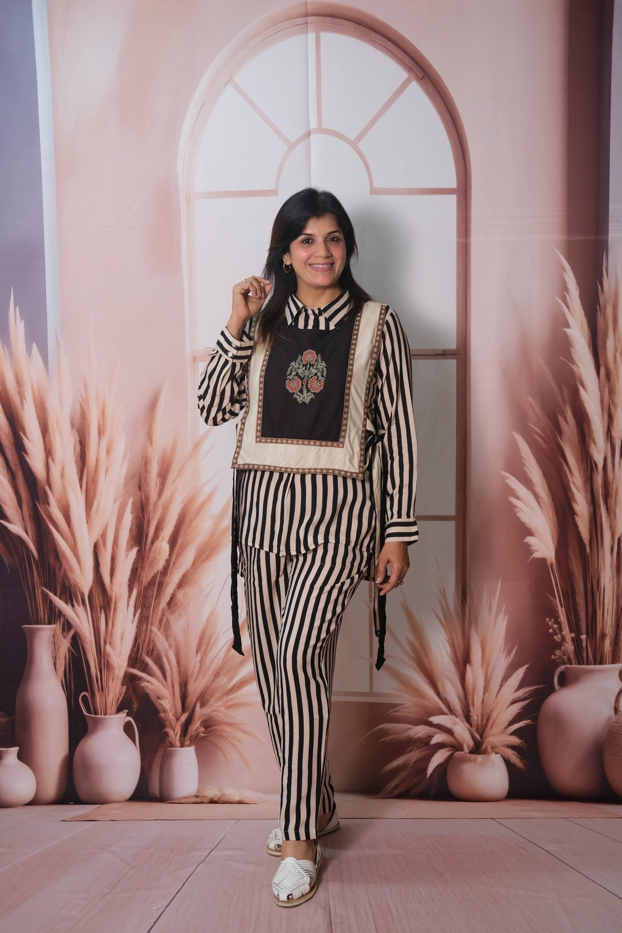 Woman posing in a stylish outfit against a decorative wall with plants and vases.