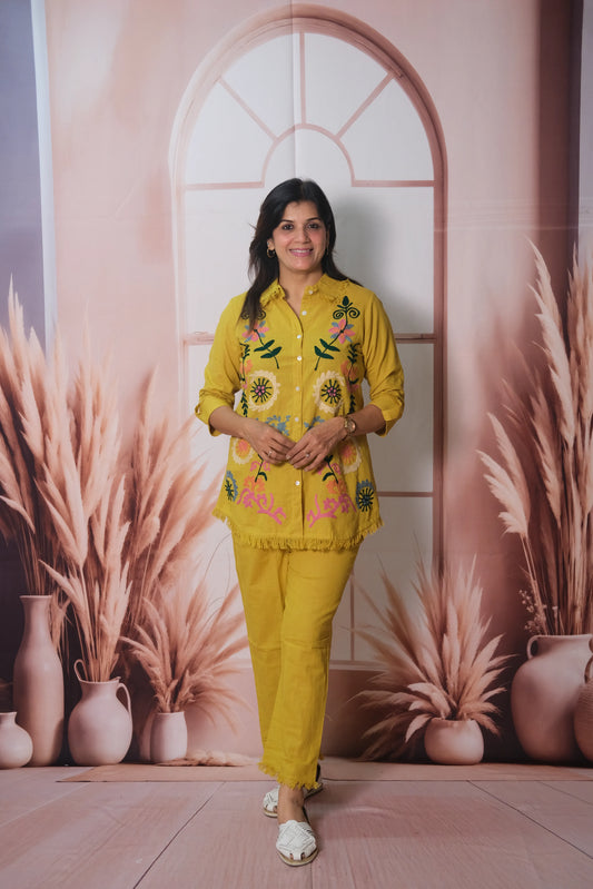 Woman in a yellow embroidered outfit standing in front of a decorative wall with plants and vases.