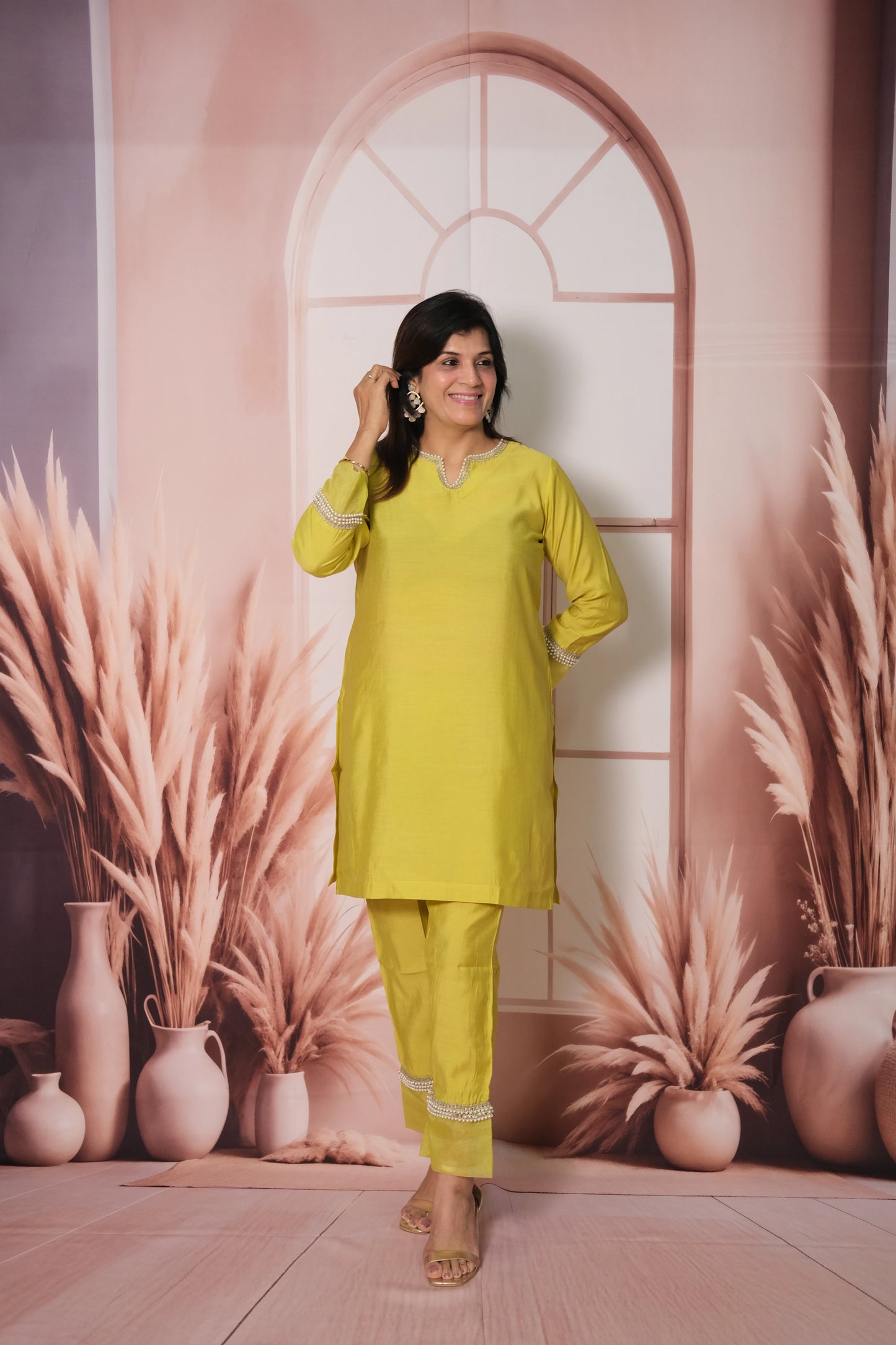 Woman in a yellow outfit standing against a decorative wall with plants and vases.