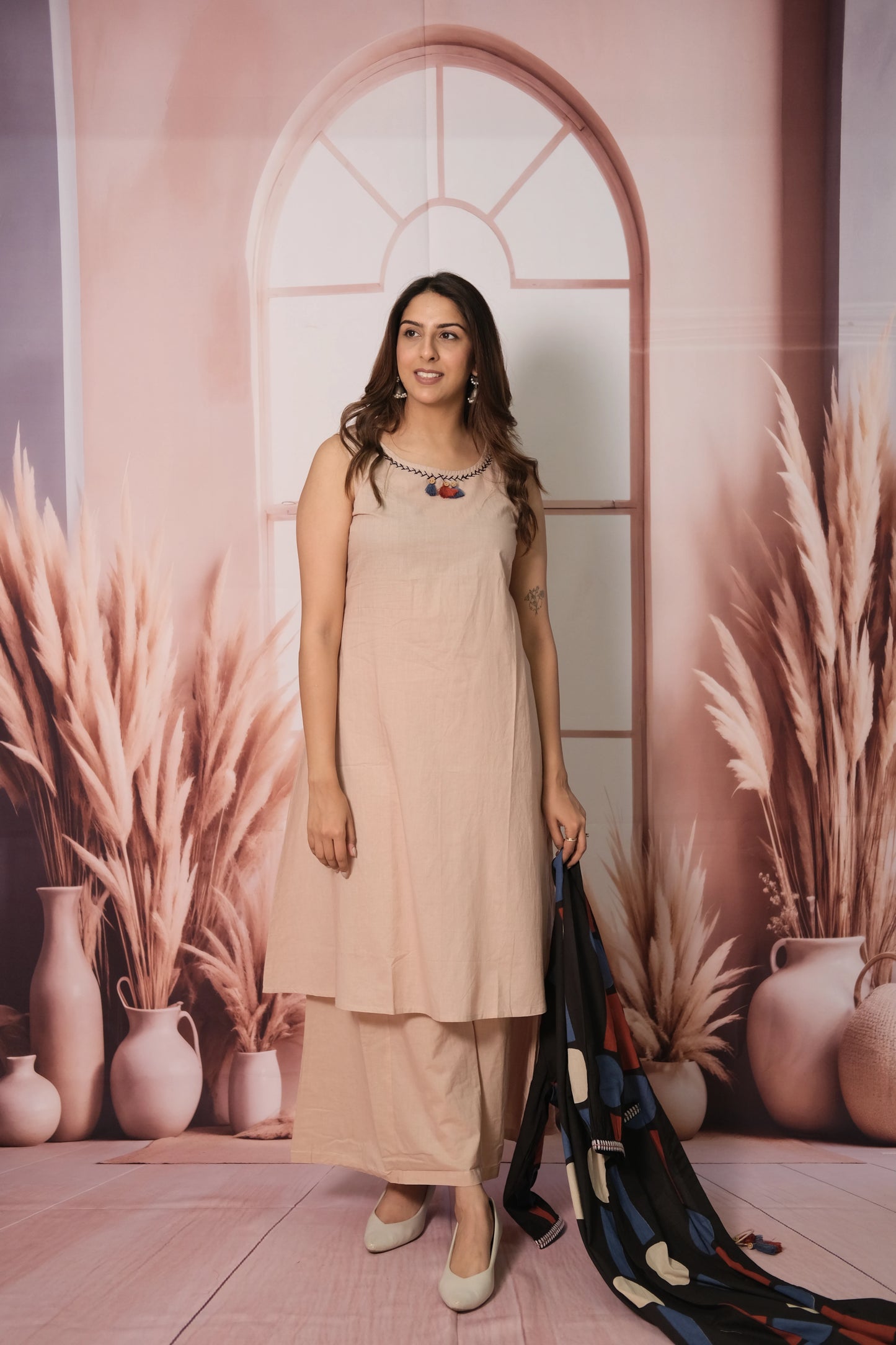 Woman in a beige dress standing in front of a decorative wall with arches and plants.