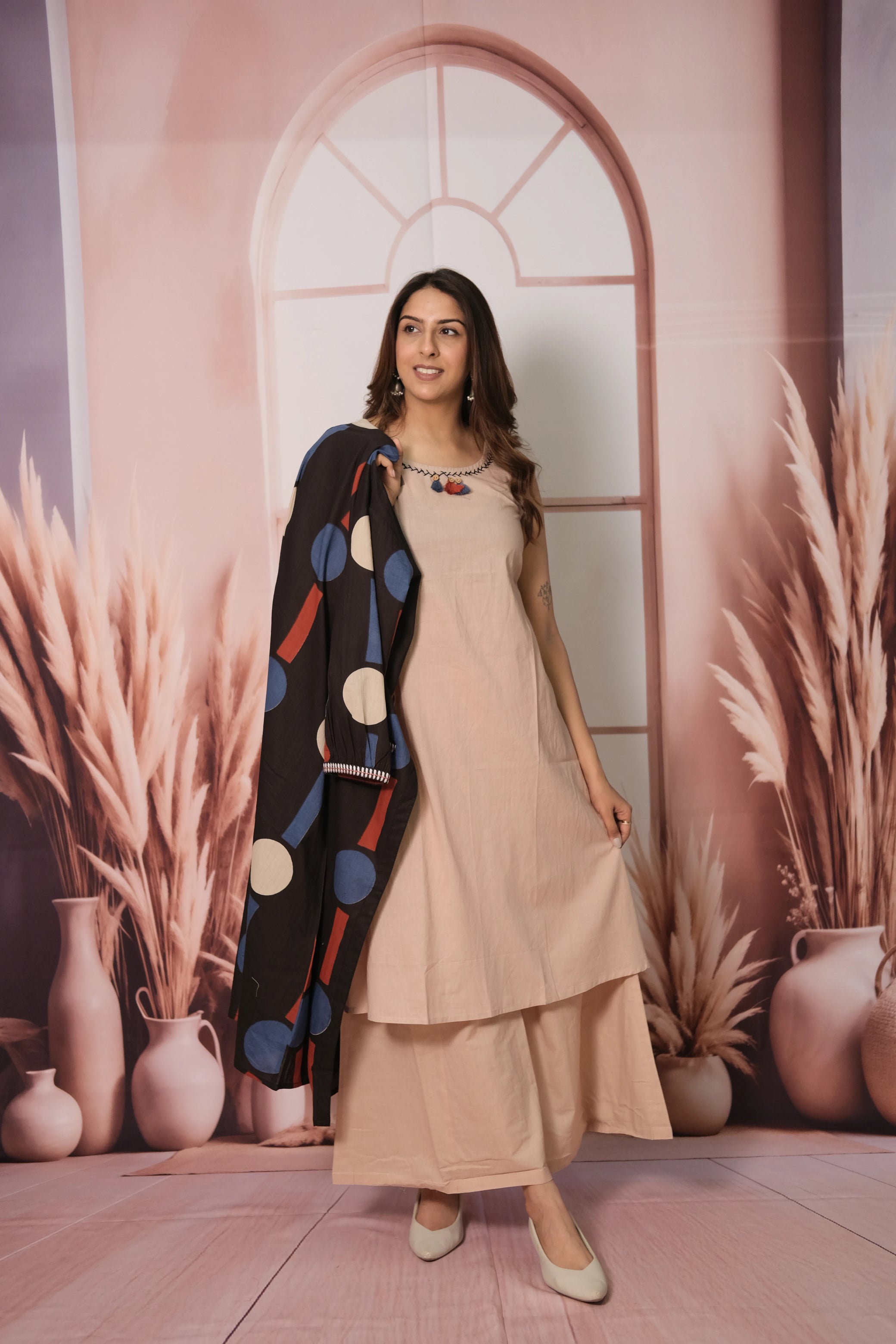 Woman in a beige outfit with a colorful cardigan standing in front of a decorative wall with arches and plants.