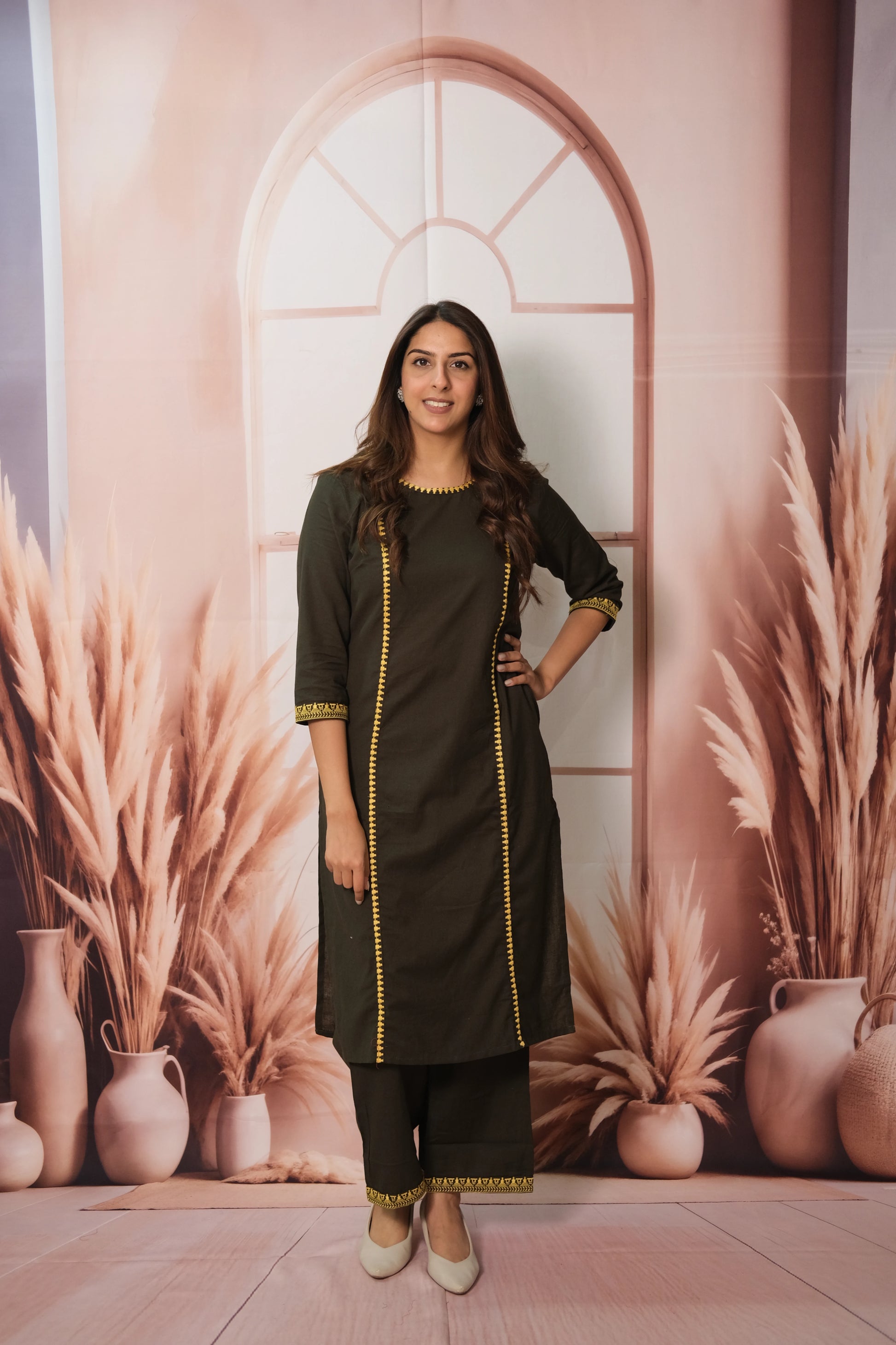 Woman in a dark green traditional outfit standing against a decorative wall with plants and vases.