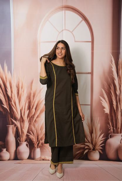 Woman in a black outfit with yellow accents standing in front of a decorative wall with arches and plants.