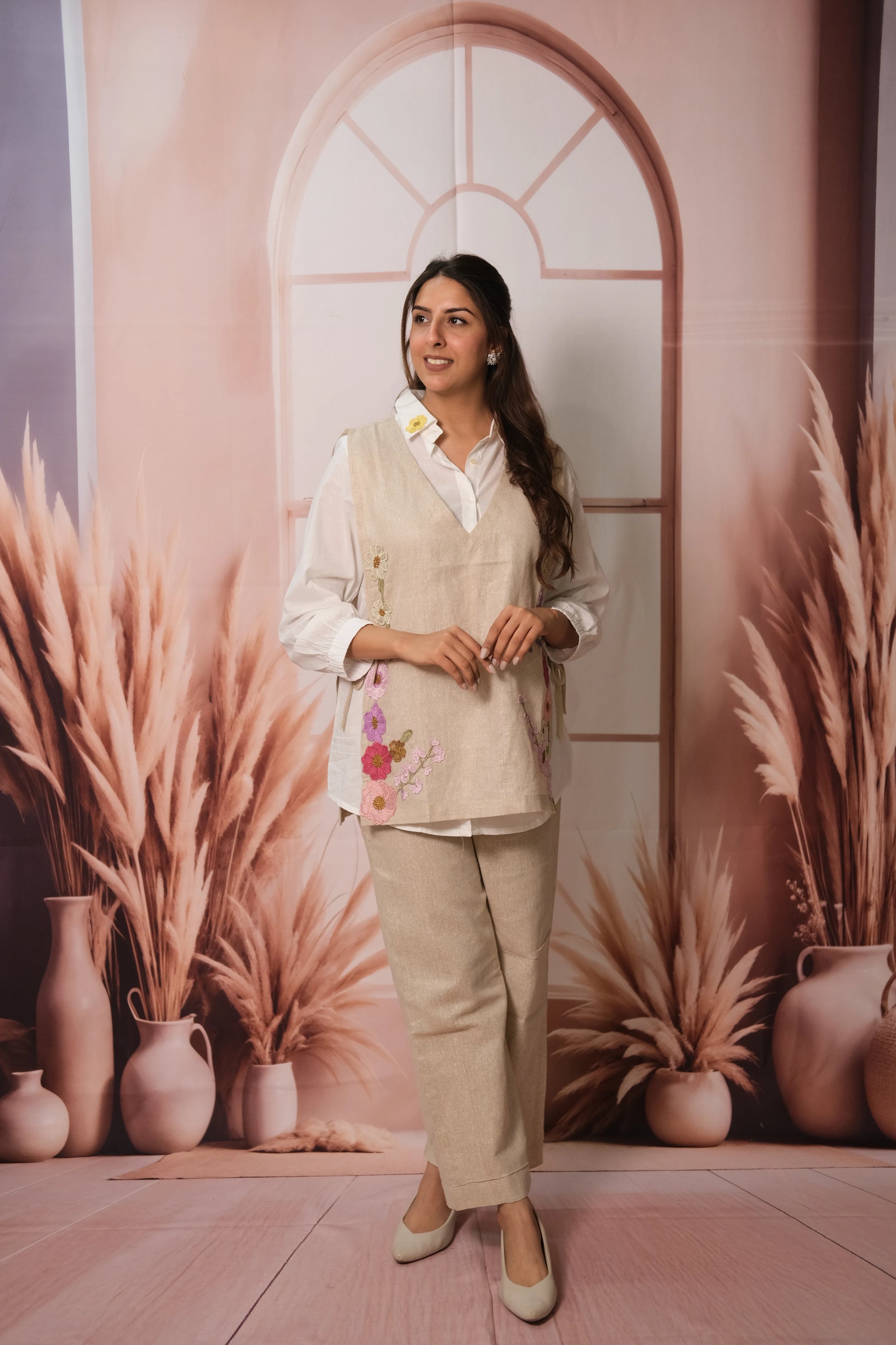 Woman in a beige outfit standing in front of a decorative wall with arches and plants.