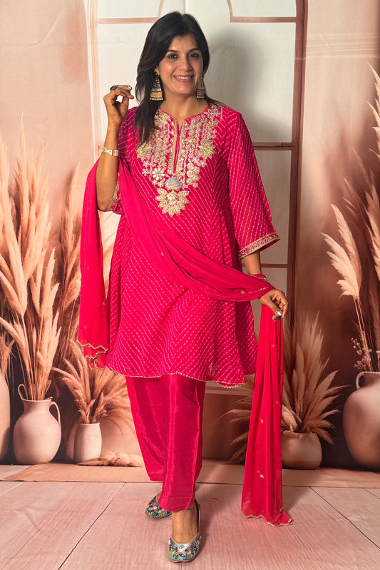 Woman in a bright pink traditional outfit with intricate designs, standing against a decorative wall.