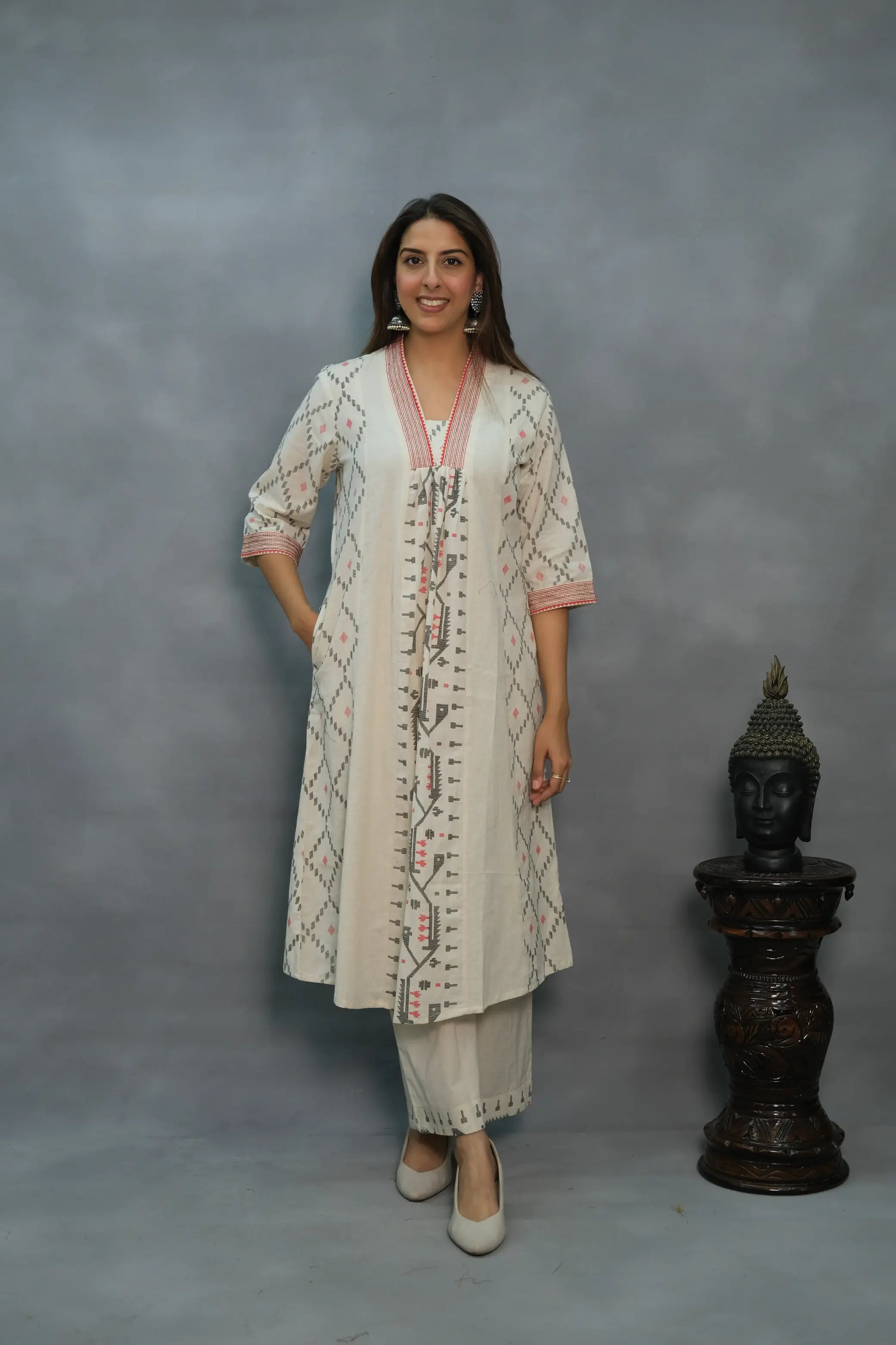 A woman modeling a white and multicolored 2-piece kurta pant set with intricate patterns, standing against a grey background with a decorative item to the side.
